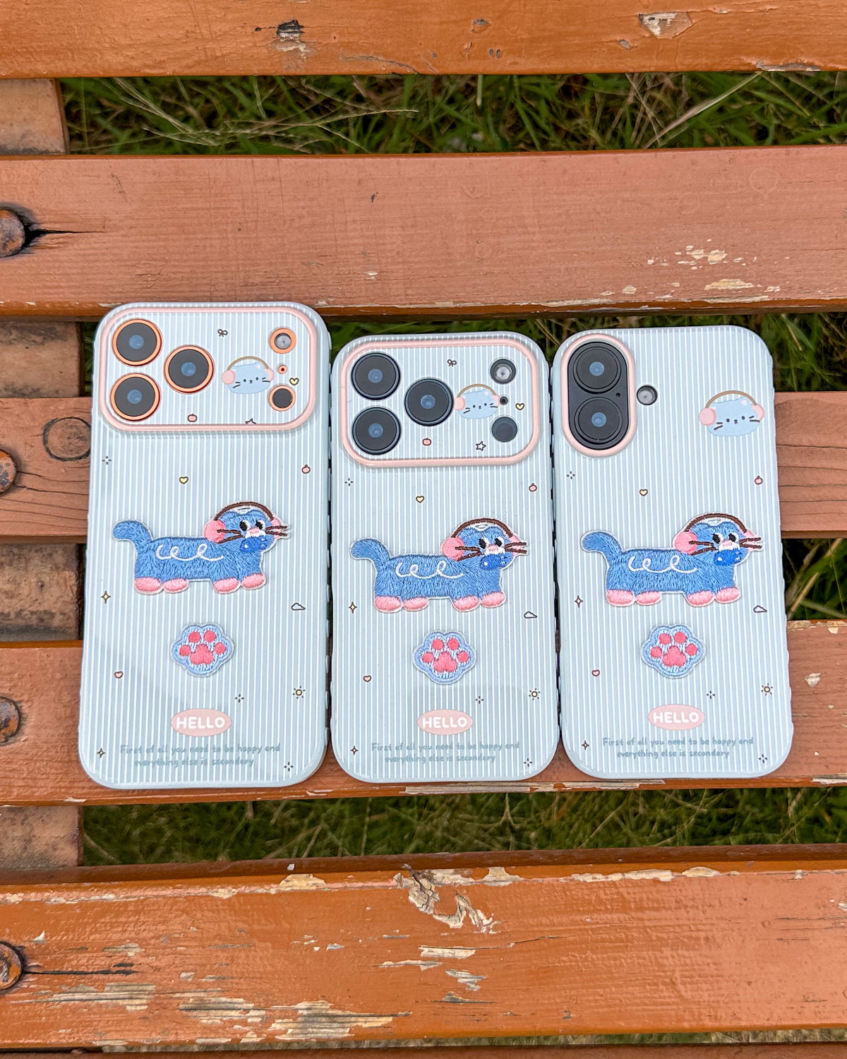 Three TOKFO Hello Cat phone cases for different iPhone models laid on a wooden bench