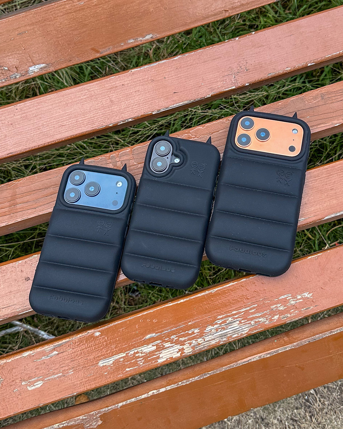 Three black TOKFO Puffer Devil Horn phone cases for different iPhone models on a wooden bench